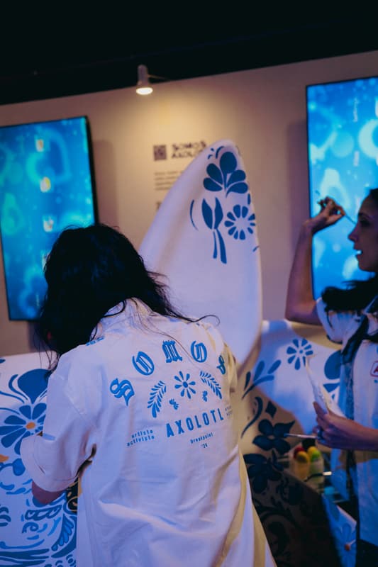 An artist in a Somos Axolotl jacket painting, display screens behind